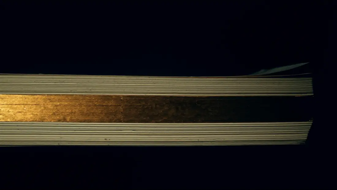 A stack of ivory pages edge-on in low light, one gilded edge catching gold